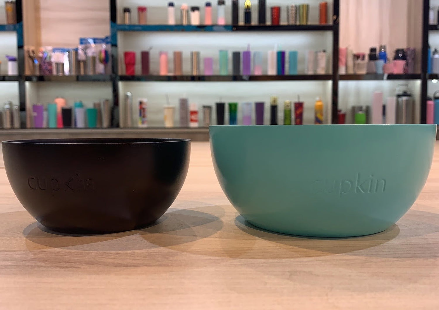 Two bowls, one black and one teal, on a wooden surface with a shelf of products in the background.
