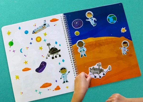 A children's spiral-bound space-themed activity book shown open on a teal surface, with a child's hands placing a sticker of a rover onto the right page. The left page has white space illustrations, and the right page shows a blue outer-space background with planets, astronauts, and an orange ground.