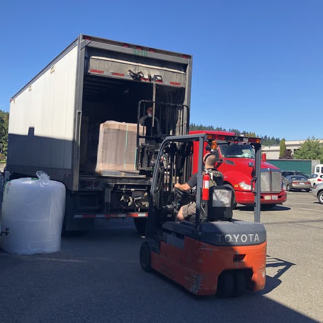 Forklift loading a large container into a truck with clear blue sky.