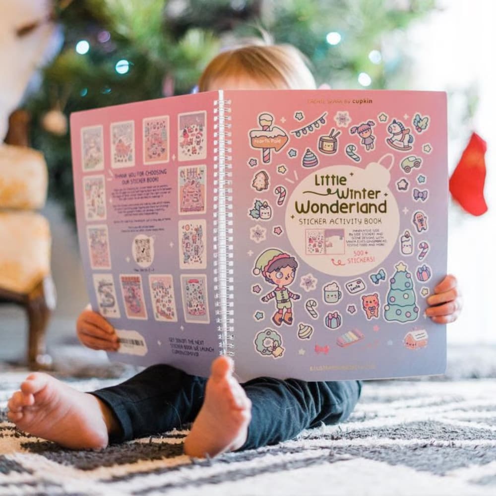 Child holding a 'Little Winter Wonderland' sticker activity book with Christmas decorations in the background.